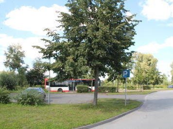 Im Vordergrund ist ein Teil der Zuwegung zum Parkplatz zu erkennen. Ein PKW parkt. Ein Baum wirft Schatten auf den Platz.
